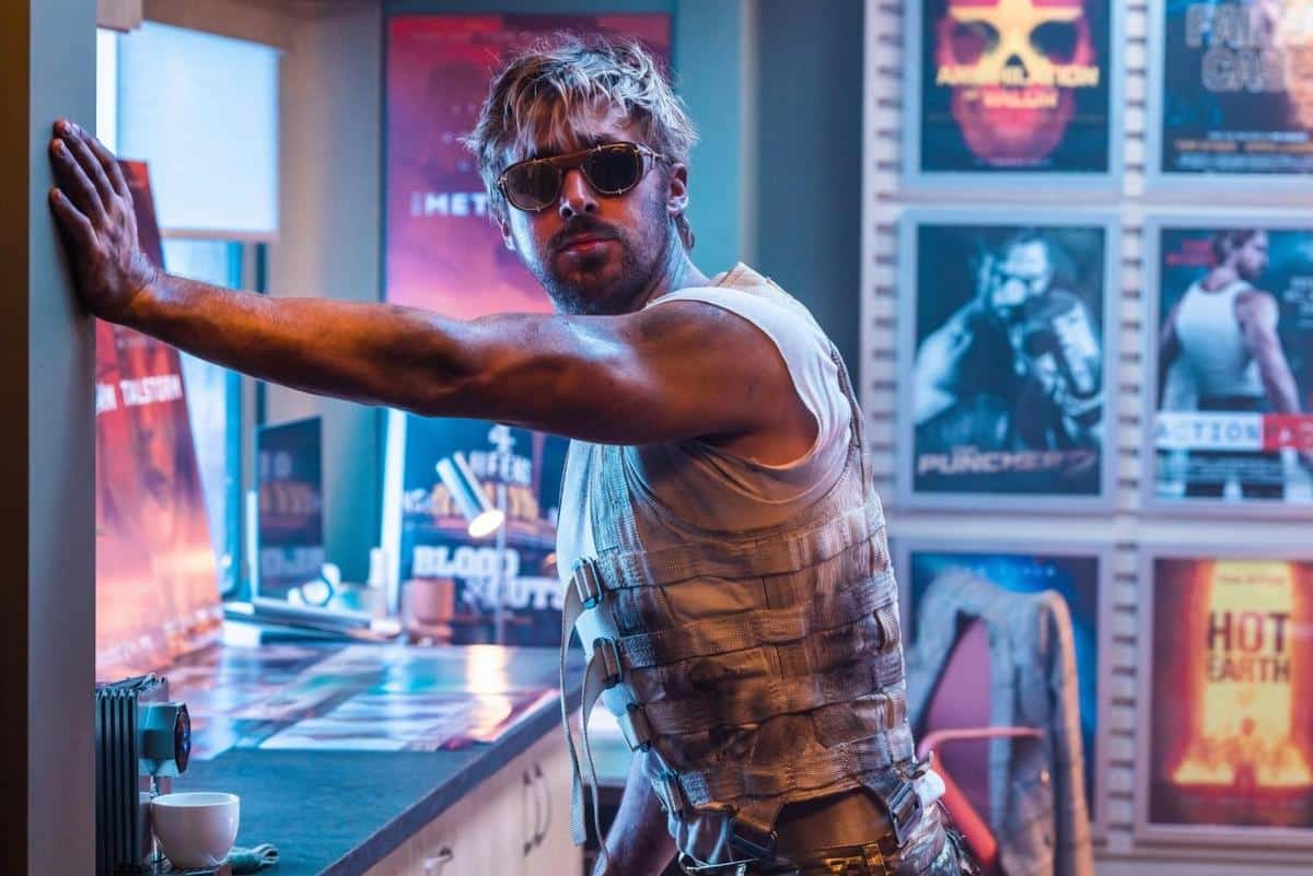 A man with tousled blond hair and sunglasses poses in a sleeveless top and tactical vest in a room filled with movie posters and neon signs, exuding a rugged, action-packed vibe.