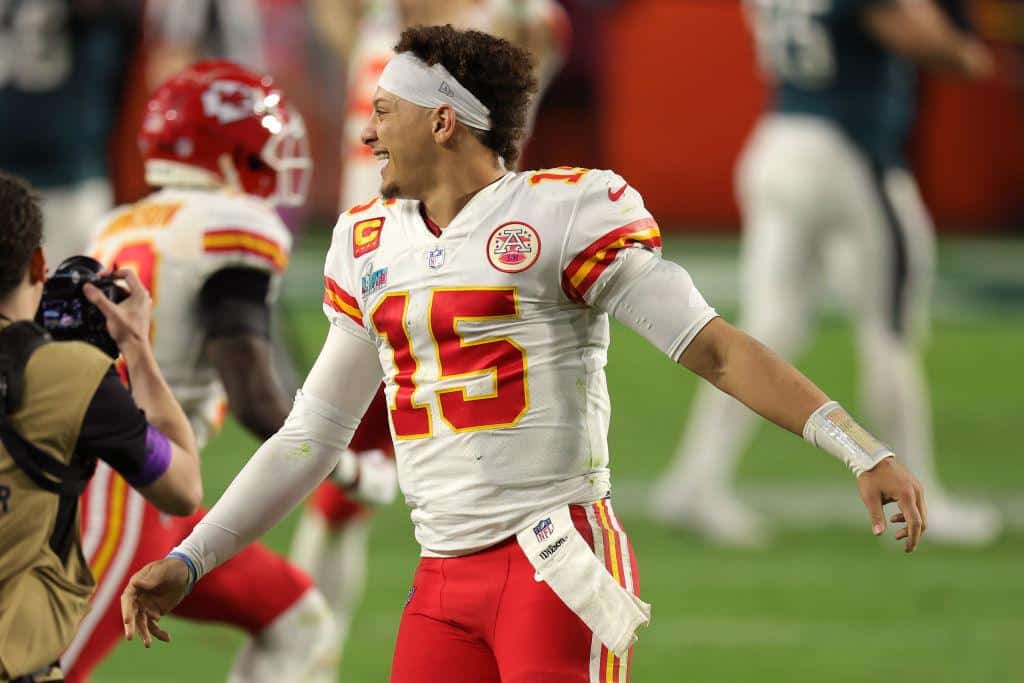 A kansas city chiefs quarterback wearing jersey number 15 celebrates on the football field. he is expressing joy, extending his right arm, while photographers capture the moment, including one in a black jacket on the left.