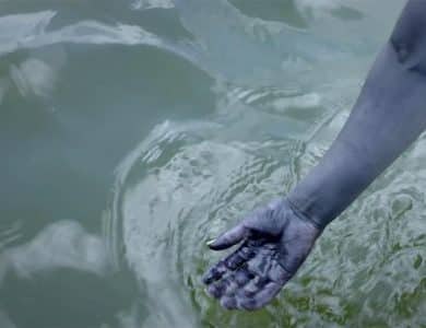 A human hand partially submerged in rippling water, reflecting a blurred image of the sky and trees. the hand appears to have streaks of black paint, suggesting a creative or artistic context.