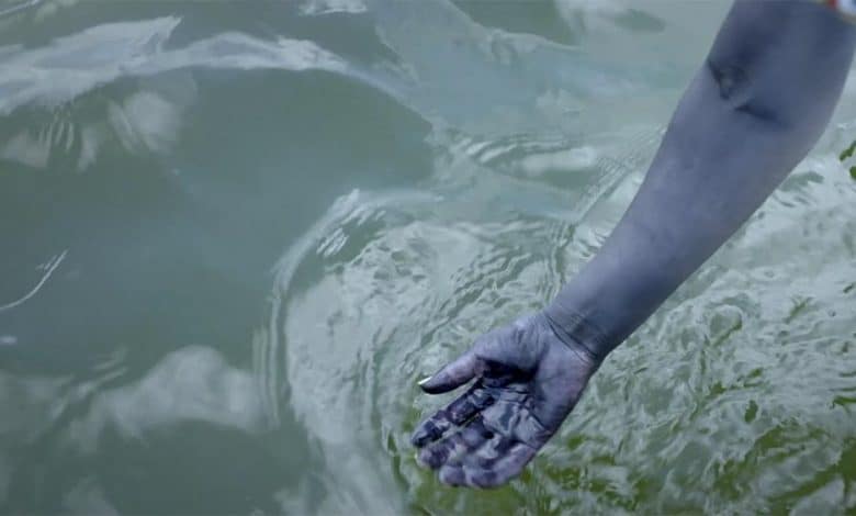 A human hand partially submerged in rippling water, reflecting a blurred image of the sky and trees. the hand appears to have streaks of black paint, suggesting a creative or artistic context.