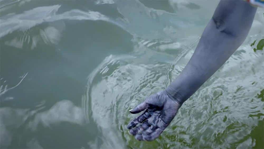 A human hand partially submerged in rippling water, reflecting a blurred image of the sky and trees. the hand appears to have streaks of black paint, suggesting a creative or artistic context.