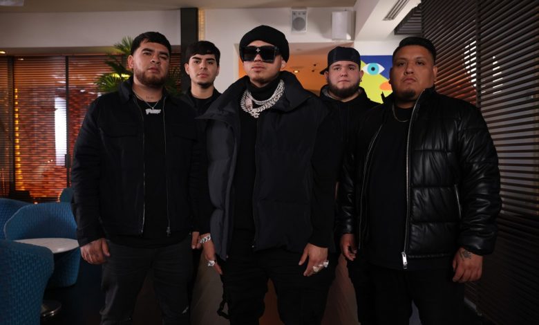 Five men stand in a lounge setting, each dressed in dark winter clothing, including jackets and hats. They are positioned in a straight line, collectively exuding a serious demeanor. The decor includes