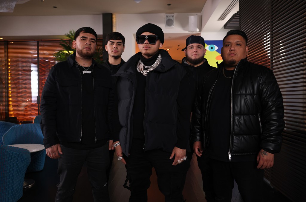 Five men stand in a lounge setting, each dressed in dark winter clothing, including jackets and hats. They are positioned in a straight line, collectively exuding a serious demeanor. The decor includes