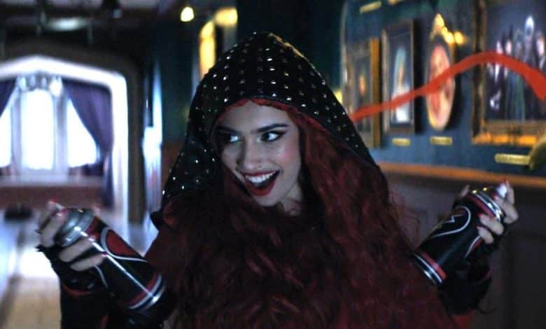 A woman with vibrant red hair and red lipstick smiles broadly, wearing a hood and holding spray paint cans with 'Red Rise' streaks in the air, inside a dimly lit hallway with framed pictures