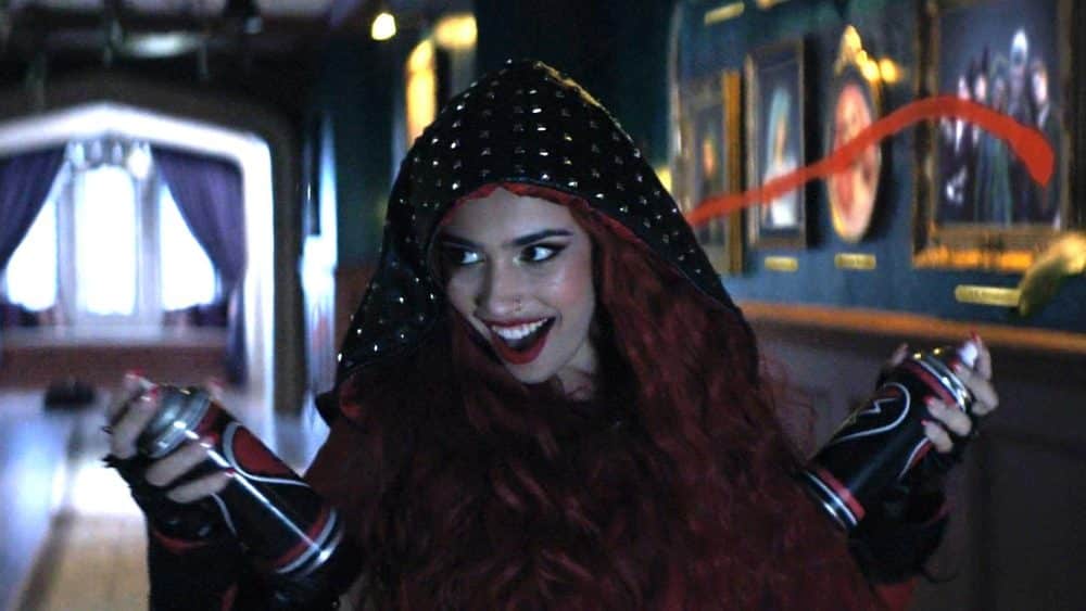 A woman with vibrant red hair and red lipstick smiles broadly, wearing a hood and holding spray paint cans with 'Red Rise' streaks in the air, inside a dimly lit hallway with framed pictures