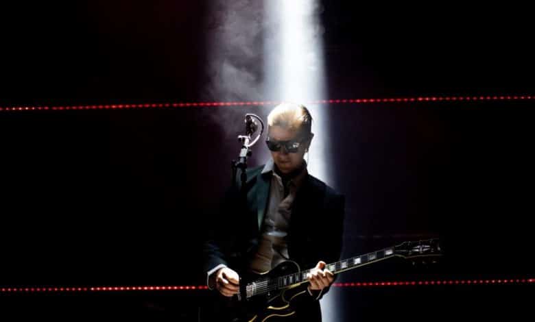 A musician in sunglasses and a suit plays an electric guitar on a dark stage, dramatically lit by a single bright spotlight from above, with hazy smoke surrounding him and red laser lights in the background during