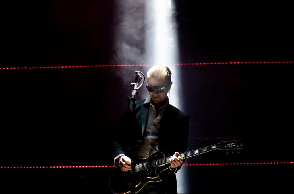 A musician in sunglasses and a suit plays an electric guitar on a dark stage, dramatically lit by a single bright spotlight from above, with hazy smoke surrounding him and red laser lights in the background during