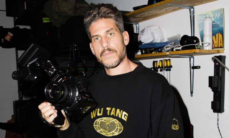 A man with short, wavy hair holds a camera, looking directly at the viewer. He wears a black Wu-Tang sweatshirt and stands in a storage room with shelves containing various items and equipment