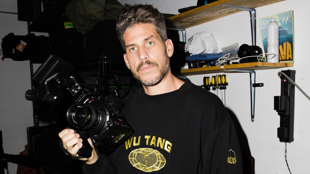 A man with short, wavy hair holds a camera, looking directly at the viewer. He wears a black Wu-Tang sweatshirt and stands in a storage room with shelves containing various items and equipment