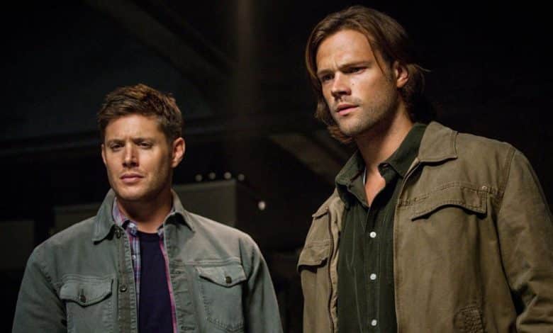 Two men stand side by side in a dimly lit room, looking intently to their left. Jared Padalecki, on the left, wears a blue jacket, and the taller man on the
