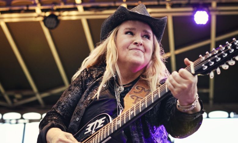 A woman in a black cowboy hat and sparkling jacket plays an elaborate guitar on stage, intensely focused with a joyful expression, surrounded by purple stage lighting, featured in Paramount+'s "I'm Not Broken