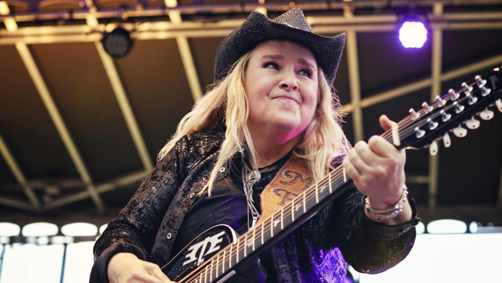 A woman in a black cowboy hat and sparkling jacket plays an elaborate guitar on stage, intensely focused with a joyful expression, surrounded by purple stage lighting, featured in Paramount+'s "I'm Not Broken
