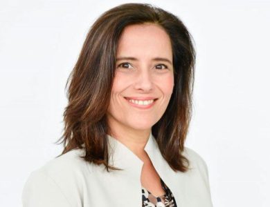 A professional headshot of a smiling woman with shoulder-length brown hair, wearing a light beige blazer and a patterned blouse, against a simple white background, captures the startling exit of Sundance CEO