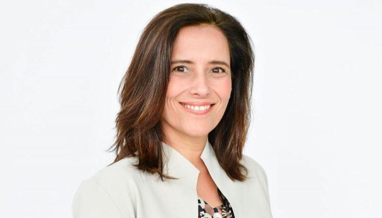 A professional headshot of a smiling woman with shoulder-length brown hair, wearing a light beige blazer and a patterned blouse, against a simple white background, captures the startling exit of Sundance CEO