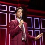 A man in a pink suit and striped tie speaks into a microphone on a stage with a red geometric patterned background. He gestures with his left hand, engaging with the audience during Netflix's "John