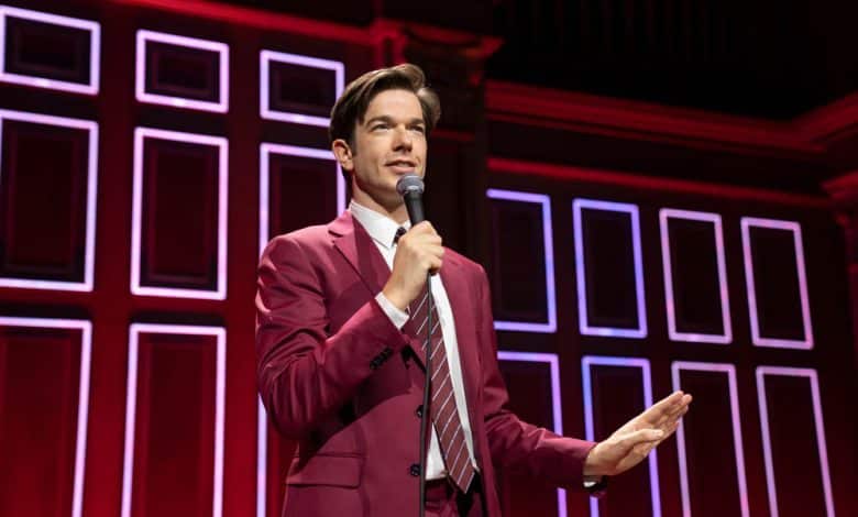 A man in a pink suit and striped tie speaks into a microphone on a stage with a red geometric patterned background. He gestures with his left hand, engaging with the audience during Netflix's "John