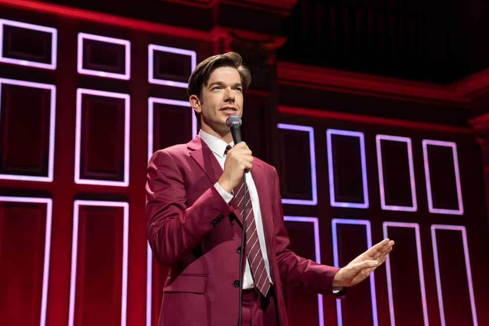 A man in a pink suit and striped tie speaks into a microphone on a stage with a red geometric patterned background. He gestures with his left hand, engaging with the audience during Netflix's "John