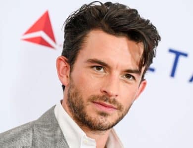 A man with dark, tousled hair and stubble gazes into the camera. He wears a light gray suit jacket over a white shirt. In the background, there's a white backdrop with red