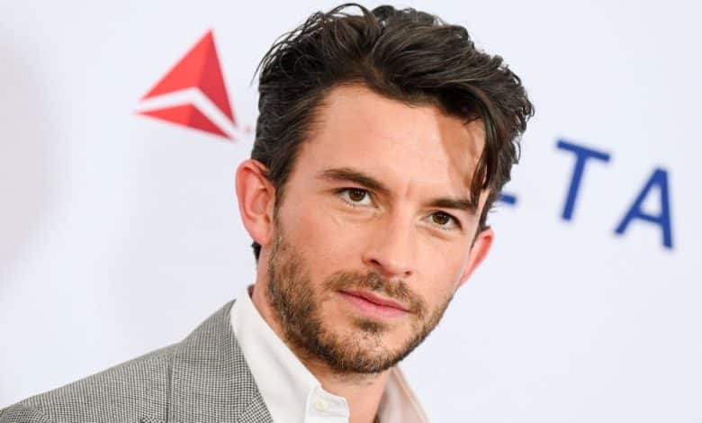 A man with dark, tousled hair and stubble gazes into the camera. He wears a light gray suit jacket over a white shirt. In the background, there's a white backdrop with red