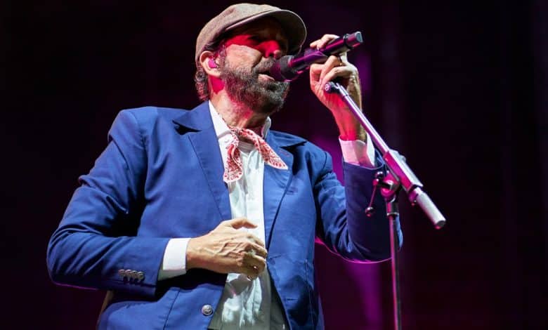 A male singer, identified as Juan Luis Guerra, performs on stage, holding a microphone close to his mouth. He wears a blue suit, white shirt, and patterned tie, complemented by