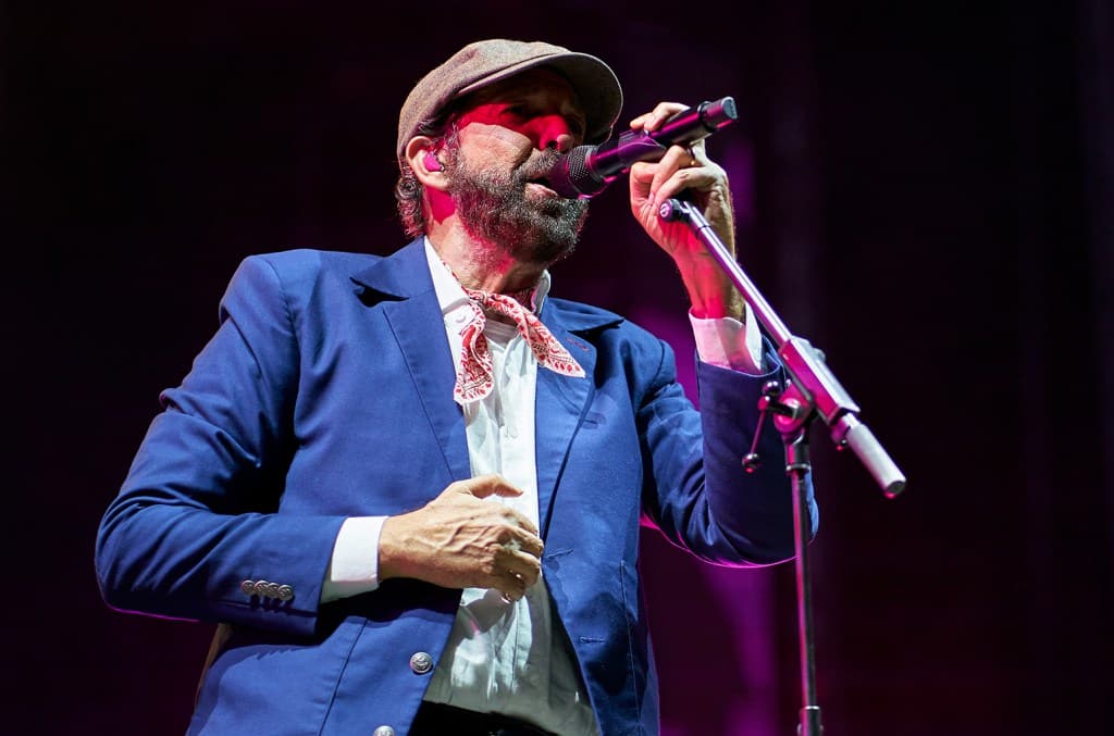 A male singer, identified as Juan Luis Guerra, performs on stage, holding a microphone close to his mouth. He wears a blue suit, white shirt, and patterned tie, complemented by