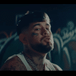 A man with tattoos on his face and neck, including a prominent cross on his forehead, sits in a dimly lit room with graffiti on the walls. He has short dark hair, a beard,