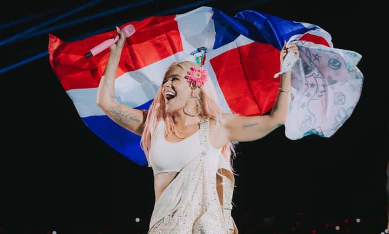 Joyful woman with pink hair, wearing a white top and a patterned skirt from Karol G's Top Fashion Moments from Mañana Será Bonito Tour, holds up the Dominican Republic flag above
