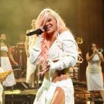 A vibrant female performer with pink hair sings passionately into a microphone on stage, adorned in a white crop top and skirt. Behind her, a band featuring a guitarist and backup singers enhances the lively musical scene