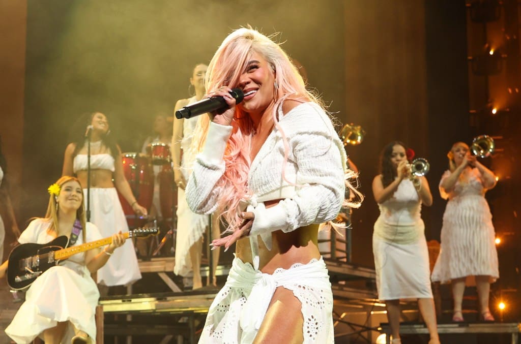 A vibrant female performer with pink hair sings passionately into a microphone on stage, adorned in a white crop top and skirt. Behind her, a band featuring a guitarist and backup singers enhances the lively musical scene