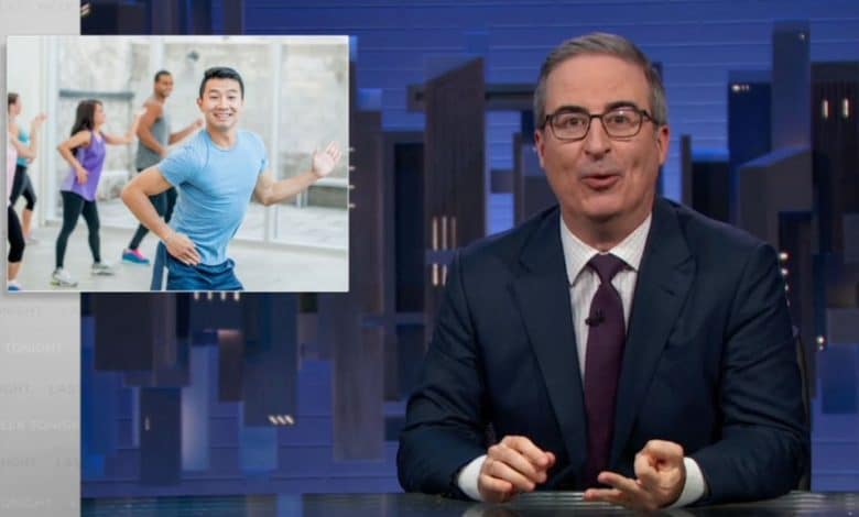 A man in a suit at a news desk smiles at the camera, with a background image on the screen showing John Oliver leading a group exercise class.