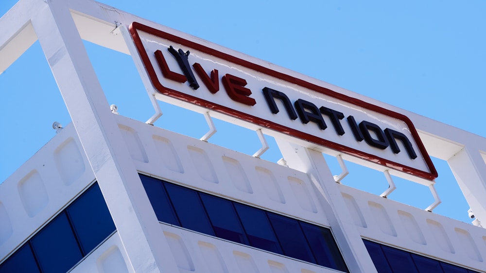 Sign reading "DOJ Sues Live Nation over Antitrust Claims" mounted on a white building structure with unique geometric patterns, set against a clear blue sky.