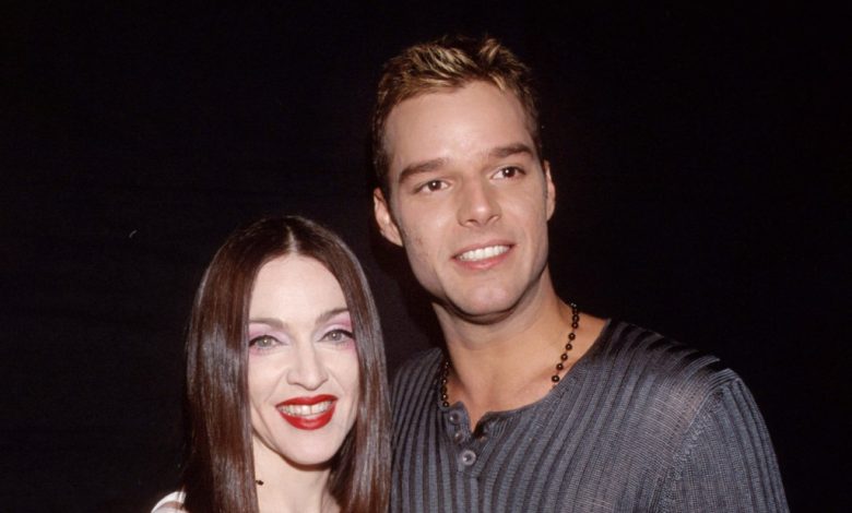 A man and a woman smile at the camera, standing close together. The woman wears bright red lipstick and a dark blouse, resembling Madonna, while the man is in a gray striped sweater and sports a