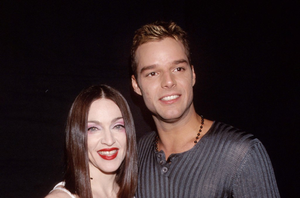A man and a woman smile at the camera, standing close together. The woman wears bright red lipstick and a dark blouse, resembling Madonna, while the man is in a gray striped sweater and sports a