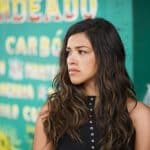 Gina Rodriguez, with long wavy hair and wearing a black sleeveless top, looks to her left with a thoughtful expression against a colorful backdrop featuring written Spanish menu items.