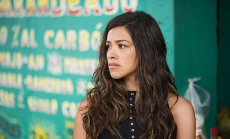 Gina Rodriguez, with long wavy hair and wearing a black sleeveless top, looks to her left with a thoughtful expression against a colorful backdrop featuring written Spanish menu items.