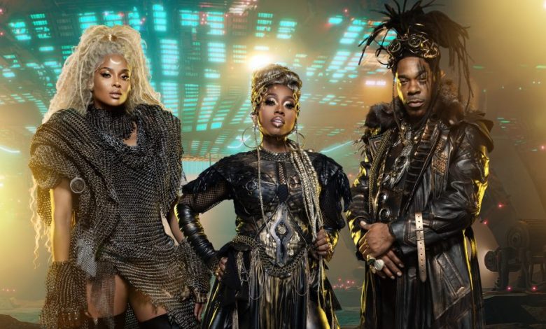 Three people pose confidently in futuristic tribal costumes against a backdrop of glowing electronic panels. The left person has voluminous curly hair and silvery attire resembling something from Missy Elliott's 1st Headline