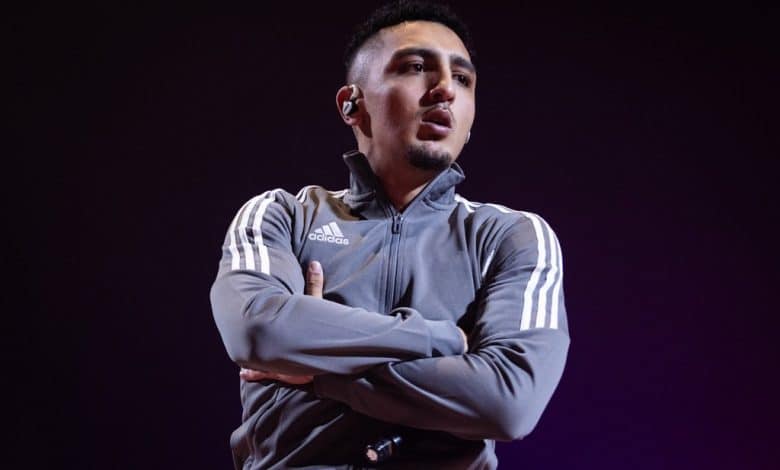 A man wearing an adidas tracksuit stands with his arms crossed, sporting a focused expression and a wireless earpiece against a dimly lit violet background as he begins his 6-month sentence for traffic offenses