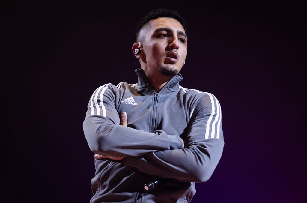 A man wearing an adidas tracksuit stands with his arms crossed, sporting a focused expression and a wireless earpiece against a dimly lit violet background as he begins his 6-month sentence for traffic offenses
