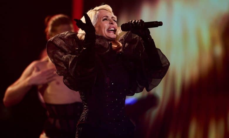 A female singer performs passionately on stage, wearing a dramatic black outfit with voluminous sleeves. A blurred dancer, part of Nebulossa's collab with Gloria Trevi, is visible behind her.