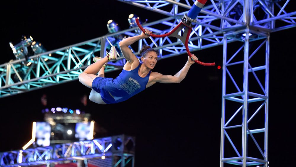 A female trapeze artist, wearing a blue costume, performs a dramatic aerial maneuver high above the ground. She is gripping red trapeze bars, surrounded by a metal framework, with a Ninja