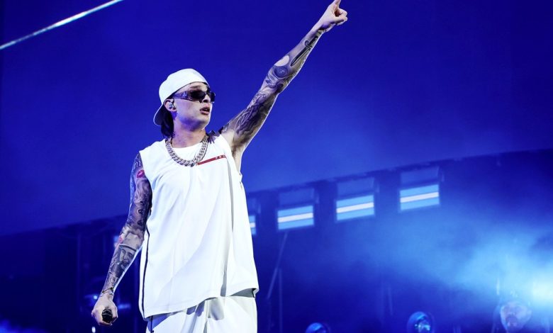 A male performer on stage, dressed in a white sleeveless top, white headband, and sunglasses, pointing upwards. He has tattooed arms, wearing a necklace associated with $1B+ Latin