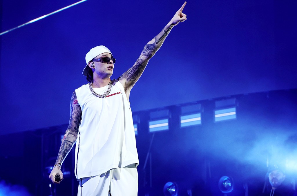 A male performer on stage, dressed in a white sleeveless top, white headband, and sunglasses, pointing upwards. He has tattooed arms, wearing a necklace associated with $1B+ Latin
