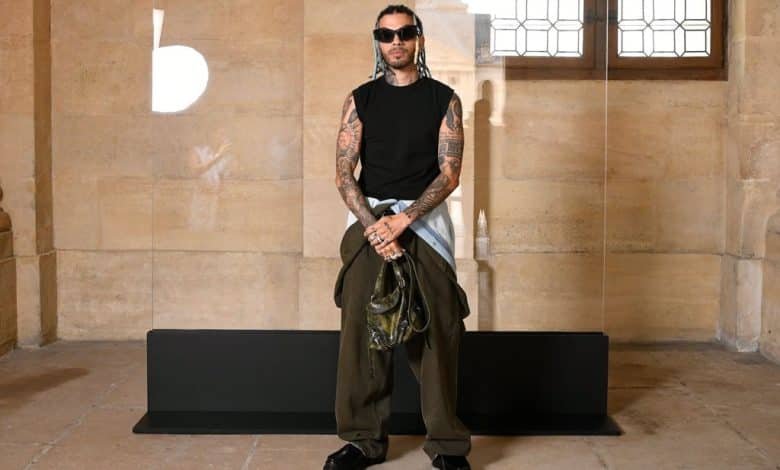 A man with tattoos stands confidently in an ornate room, wearing a sleeveless black top, baggy black pants, and holding a green jacket. His hair is styled upwards, and he has a