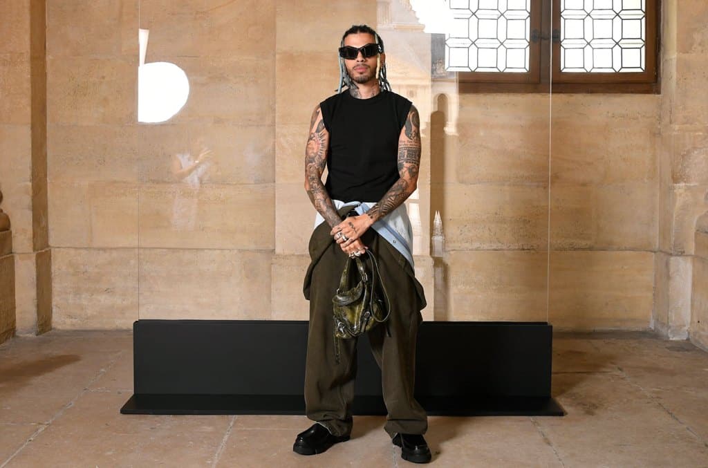 A man with tattoos stands confidently in an ornate room, wearing a sleeveless black top, baggy black pants, and holding a green jacket. His hair is styled upwards, and he has a
