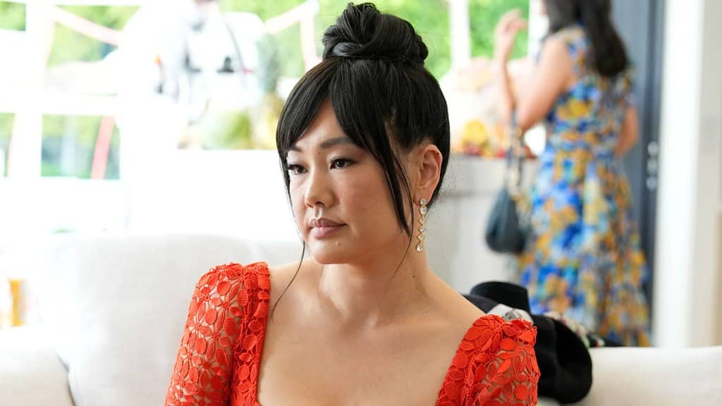 Crystal Kung Minkoff, with a top bun hairstyle, wearing a red lace dress and dangling earrings, sits thoughtfully on a sofa in a bright room with blurred figures in the background.