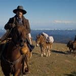 A man in a cowboy hat and coat rides a brown horse in a vast, sunny landscape with distant snow-covered mountains, in Kevin Costner's Western epic "Horizon." Two more individuals on horses