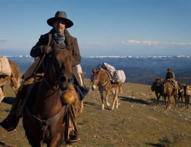 A man in a cowboy hat and coat rides a brown horse in a vast, sunny landscape with distant snow-covered mountains, in Kevin Costner's Western epic "Horizon." Two more individuals on horses