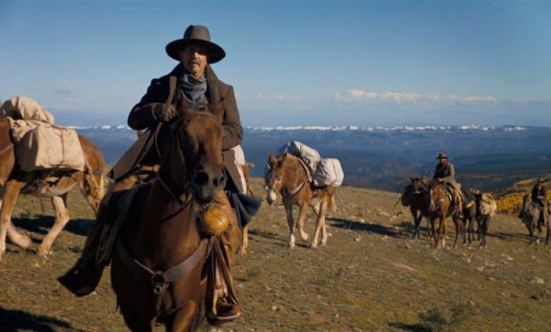 A man in a cowboy hat and coat rides a brown horse in a vast, sunny landscape with distant snow-covered mountains, in Kevin Costner's Western epic "Horizon." Two more individuals on horses