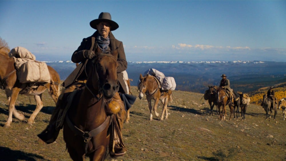 A man in a cowboy hat and coat rides a brown horse in a vast, sunny landscape with distant snow-covered mountains, in Kevin Costner's Western epic "Horizon." Two more individuals on horses
