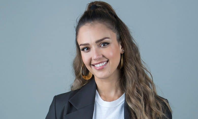 Jessica Alba, a smiling woman with long, wavy brown hair wearing large hoop earrings and a black blazer over a light top, stands against a plain gray background.